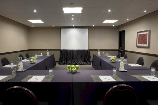 A conference room with a U-shaped table setup, featuring notepads, water glasses, carafes with lime slices, bowls of green apples, and a screen.