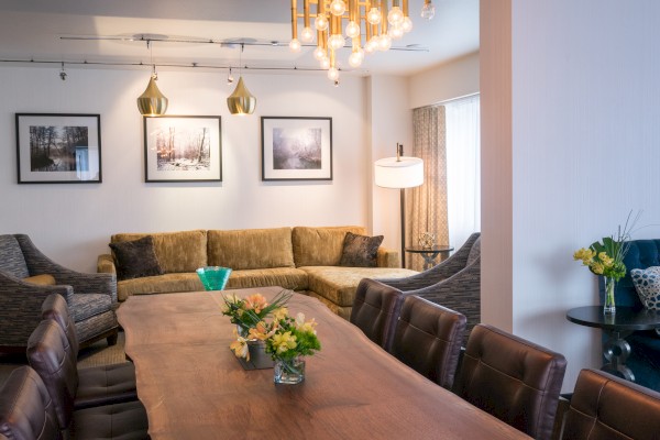 A stylish living room with a large wooden table, flowers, sectional sofa, modern lighting, and framed artwork on the walls.