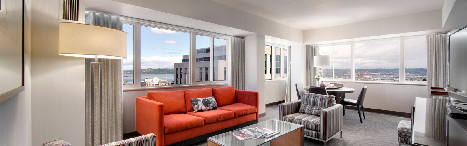 A modern, spacious living room features a red sofa, striped chairs, a glass-top coffee table, and large windows with cityscape views in the background.