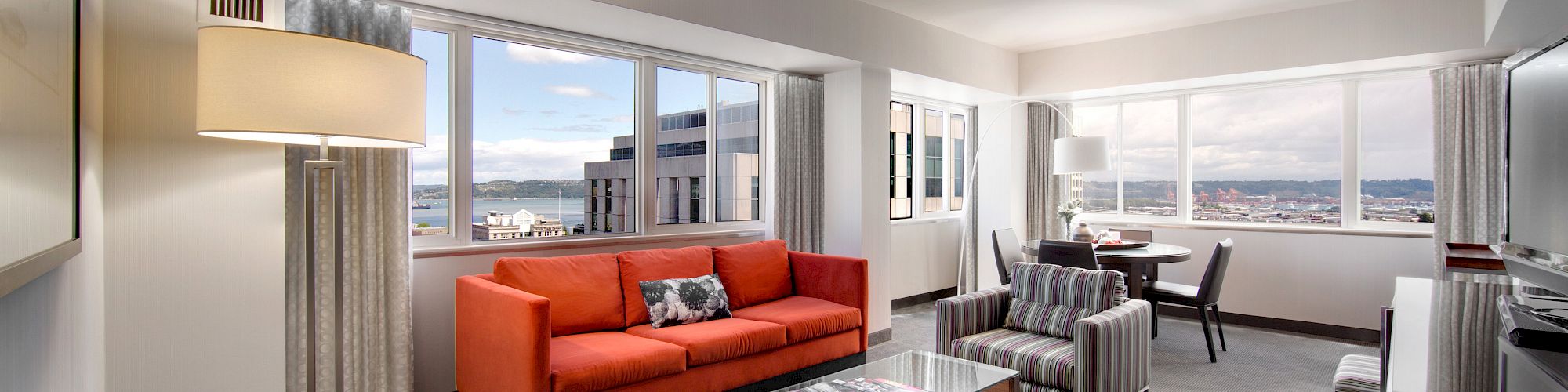 A modern, spacious living room features a red sofa, striped chairs, a glass-top coffee table, and large windows with cityscape views in the background.