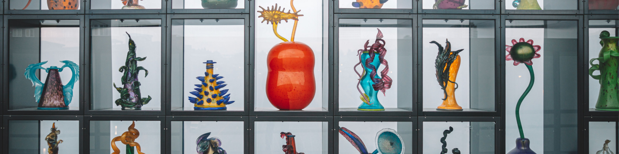 The image shows an array of artistic, colorful glass sculptures displayed in a glass cabinet, each with unique and intricate designs.