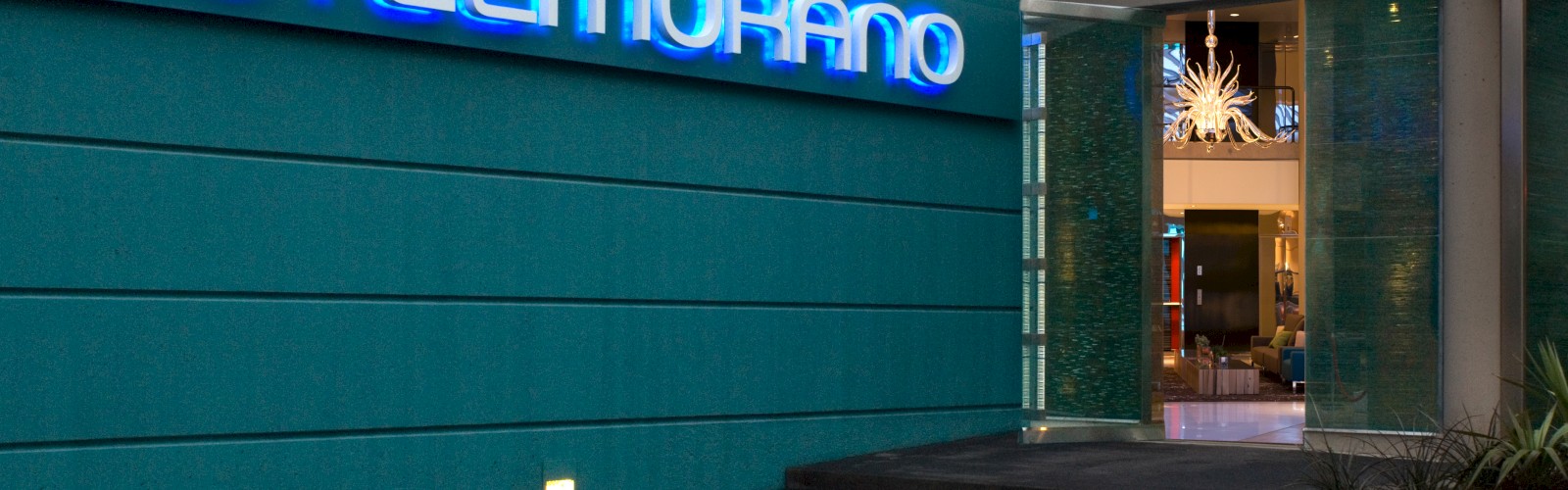 The image shows the entrance of Hotel Murano, with a blue neon sign and a modern design. The interior appears to be well-lit and stylish.