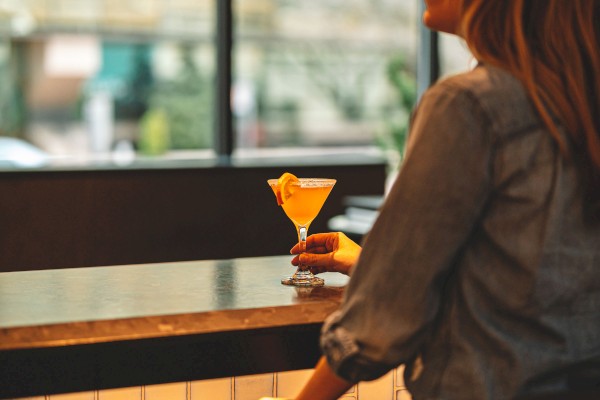 A person is sitting at a bar, holding a cocktail glass, with a relaxed expression, and a blurred view of windows in the background.