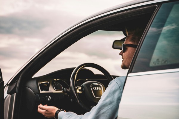 A person wearing sunglasses is sitting in a car with the door open, looking outside at a cloudy sky.