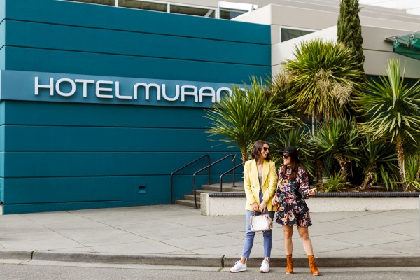 Two women stand outside the blue building of Hotel Murano, surrounded by plants, in a casual and stylish manner.