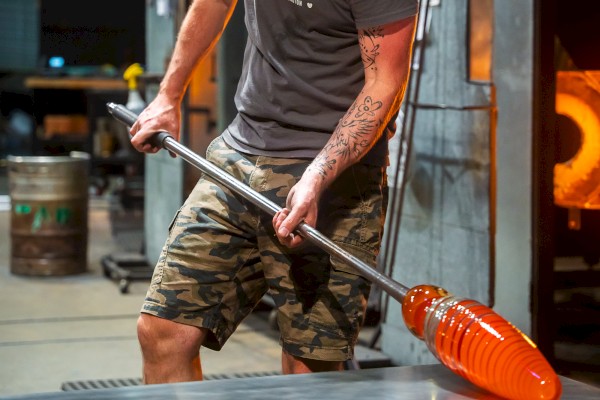 A person is glassblowing, using a metal rod to shape molten glass in a workshop, wearing a gray t-shirt and camo shorts.