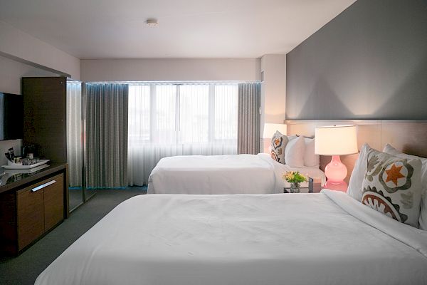 A hotel room features two beds with white linens, bedside lamps, a TV, a window with curtains, and a small kitchen area.
