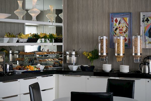 A hotel breakfast buffet area with cereal dispensers, plates, fruits, pastries, and various food items, decorated with flowers and modern art.
