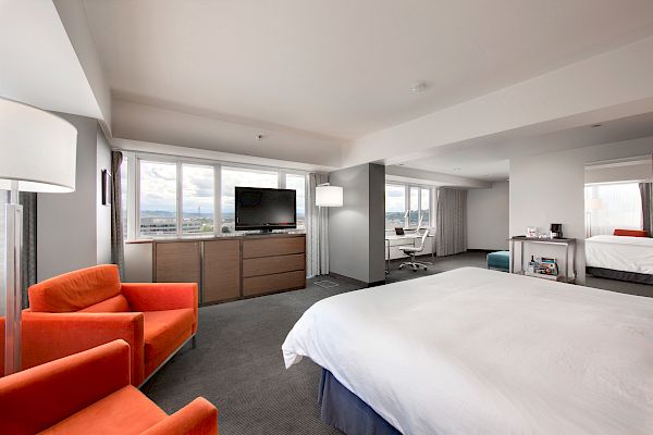 The image shows a modern hotel room with a large bed, orange chairs, a flat-screen TV, a desk, and a window offering a scenic view, ending the sentence.
