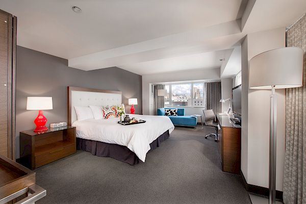 A spacious hotel room with a king-sized bed, nightstands, lamps, a sofa, a desk, a large window, and modern decor.
