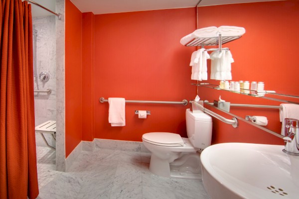 This image shows a bathroom with orange walls, a toilet, a sink, and a shower area with a red curtain. White towels are on a shelf.