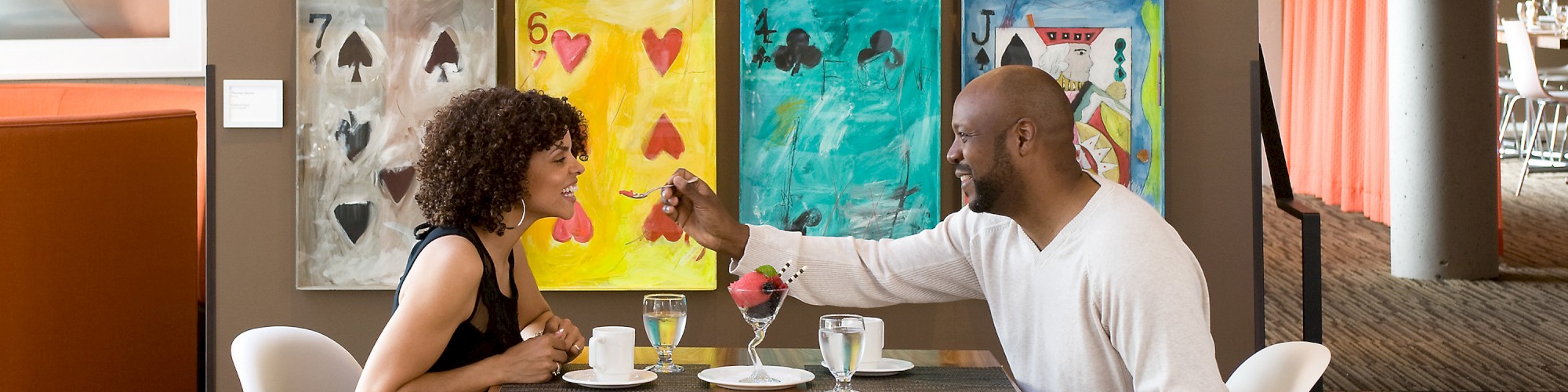 A couple sits at a table in a modern cafe with colorful card-themed art on the wall. They enjoy coffee and cake while smiling at each other.