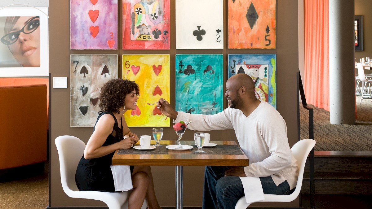 A couple is dining in a modern restaurant, surrounded by colorful card-themed artwork on the wall and a portrait photo on the left.