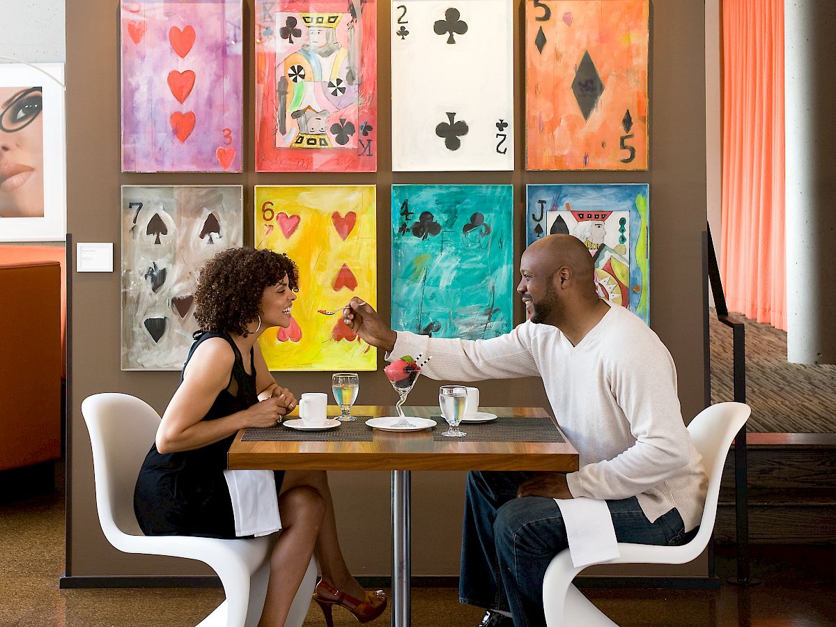 A couple is dining in a modern restaurant, surrounded by colorful card-themed artwork on the wall and a portrait photo on the left.