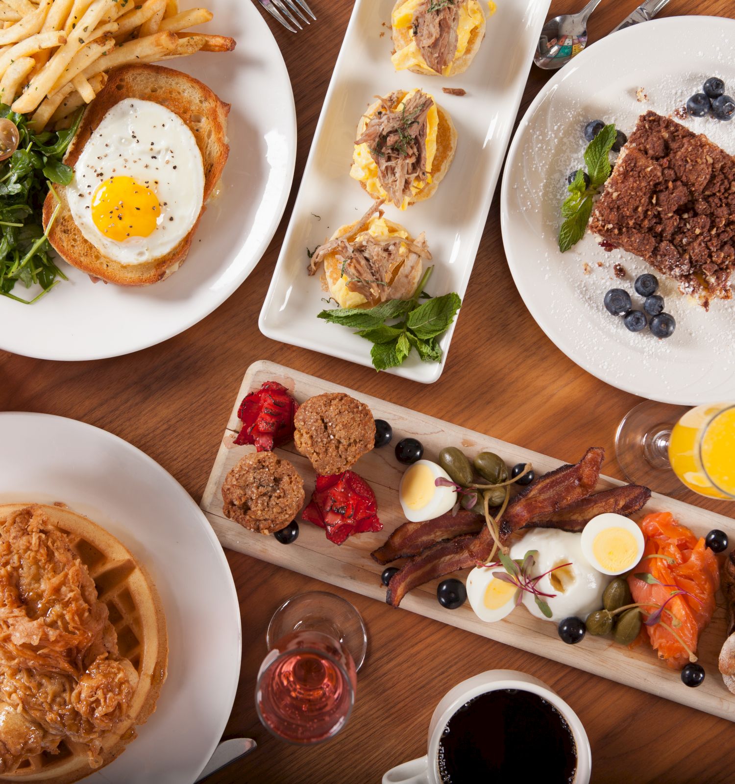 The image shows a variety of breakfast dishes on a wooden table, including waffles, salad with egg, pastries, fruit, drinks, and other assorted items.