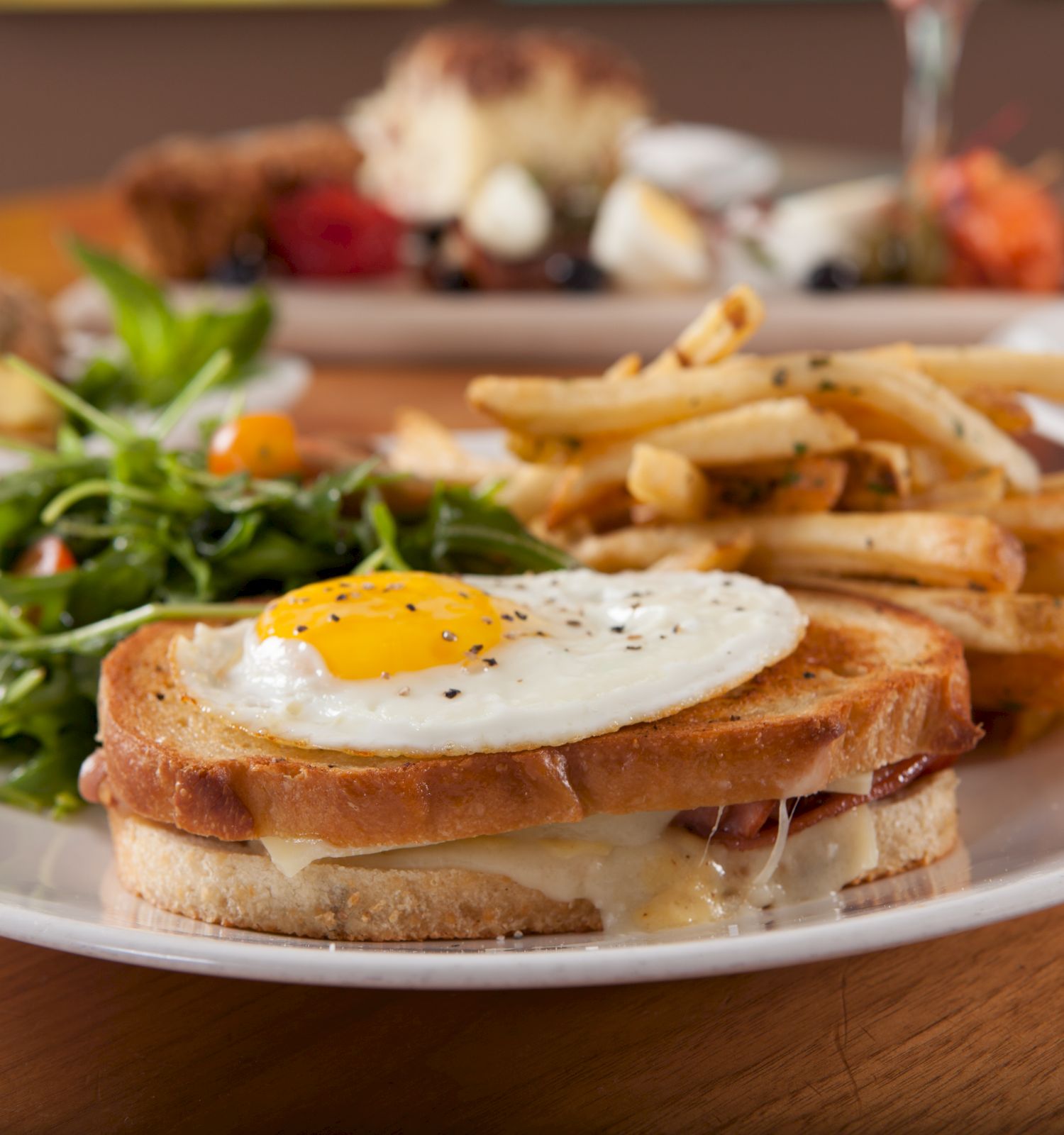 A plate featuring a fried egg on toasted sandwich, fries, and a side salad with other dishes in the background.