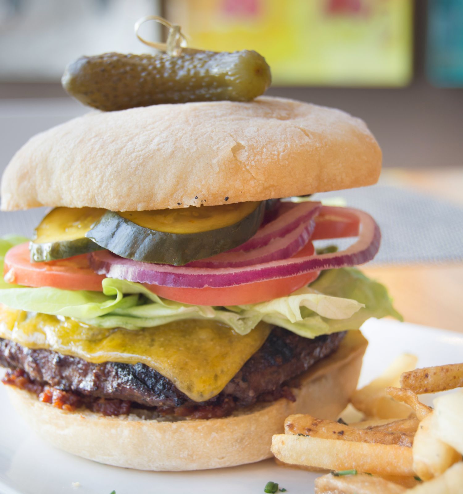 A cheeseburger with lettuce, tomato, pickles, and onion, topped with a pickle, served with fries and a drink in the background.
