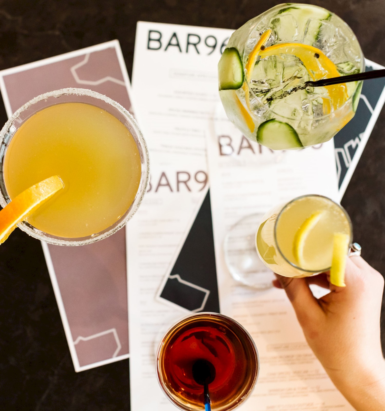 The image shows four cocktails on a table with menus from a place called 