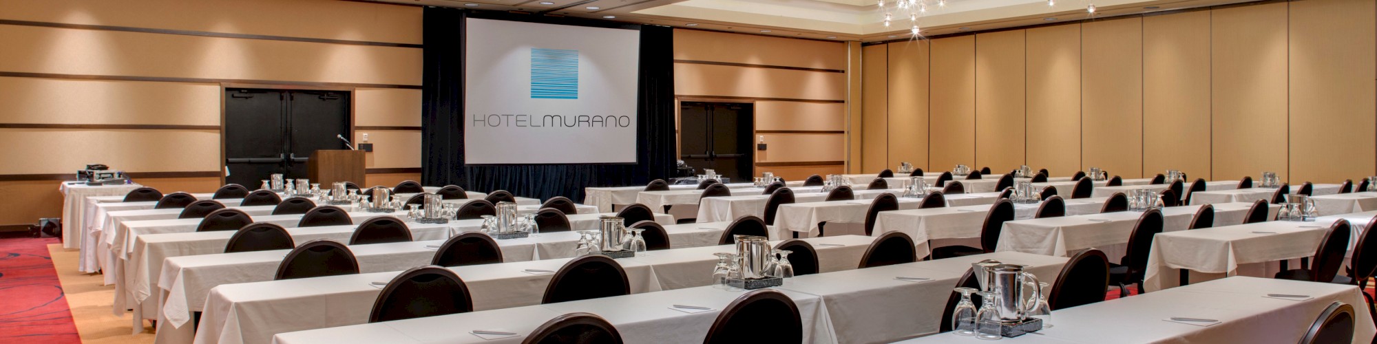 A conference room set up with long tables, chairs, and water pitchers, facing a screen with 