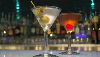 Two martini glasses, one with olives, the other with a cherry, are set against a blurred bar background with various bottles.