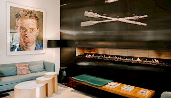 A modern living room with a sofa, art piece, and fireplace. Wooden tables and books add a cozy touch, with decorative oars above the fire.