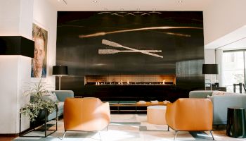A modern lounge with leather chairs, floor lamps, plants, a long fireplace, and a large abstract wall art piece, lit with natural light through windows.