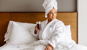 A person in a bathrobe and towel, sitting in bed holding a glass of coffee or similar beverage, looking to the side.