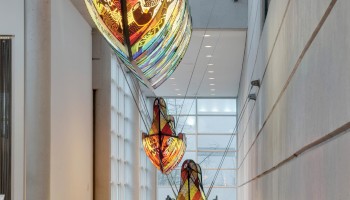 The image shows three colorful boat-like hanging sculptures inside a modern building with large windows and sleek architectural lines.