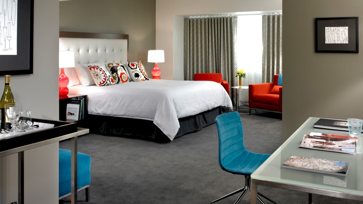 A modern hotel room with a king bed, desk with chair, orange armchair, and floor-to-ceiling window, featuring contemporary decor and artwork.