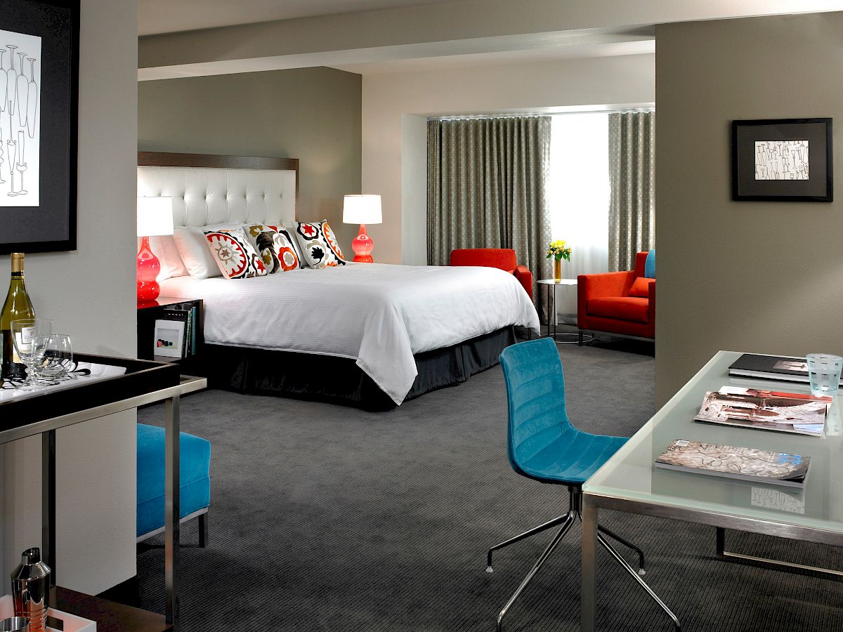A modern hotel room with a king bed, desk with chair, orange armchair, and floor-to-ceiling window, featuring contemporary decor and artwork.