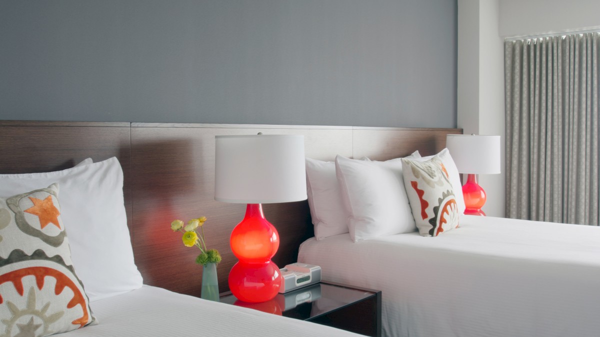 A modern bedroom with two double beds, white linens, colorful pillows, red bedside lamps, flowers, and a phone on the nightstand, gray wall.