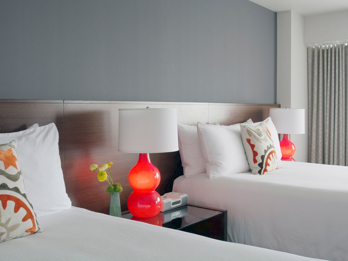 A modern bedroom with two double beds, white linens, colorful pillows, red bedside lamps, flowers, and a phone on the nightstand, gray wall.