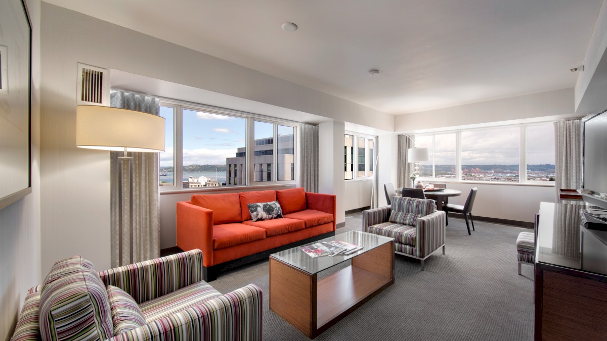 A modern living room with large windows, a red sofa, striped armchairs, a glass coffee table, a TV, and a dining area with scenic city views.