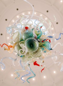 A colorful glass sculpture with intricate, swirling designs hanging from a domed ceiling with bright lights.