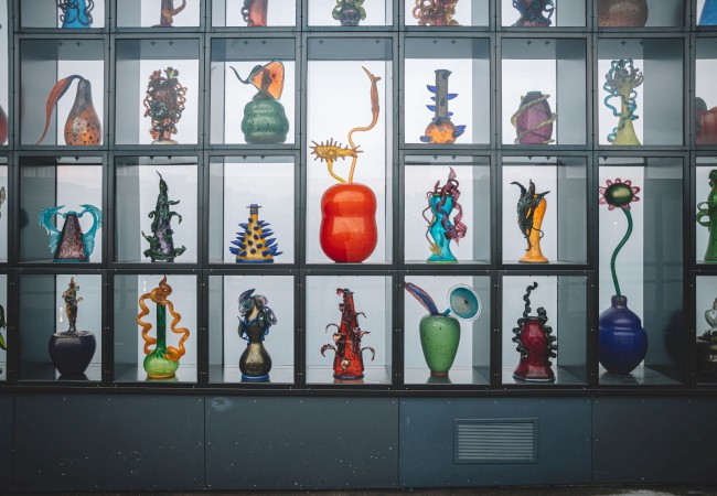 The image shows a wall display of colorful, abstract glass sculptures in various shapes and sizes, each housed in its own cubby.