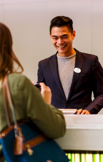 A woman is speaking to a smiling man at a reception or service counter. Both appear engaged in a friendly conversation.