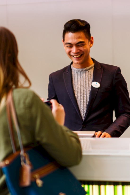 A woman is speaking to a smiling man at a reception or service counter. Both appear engaged in a friendly conversation.