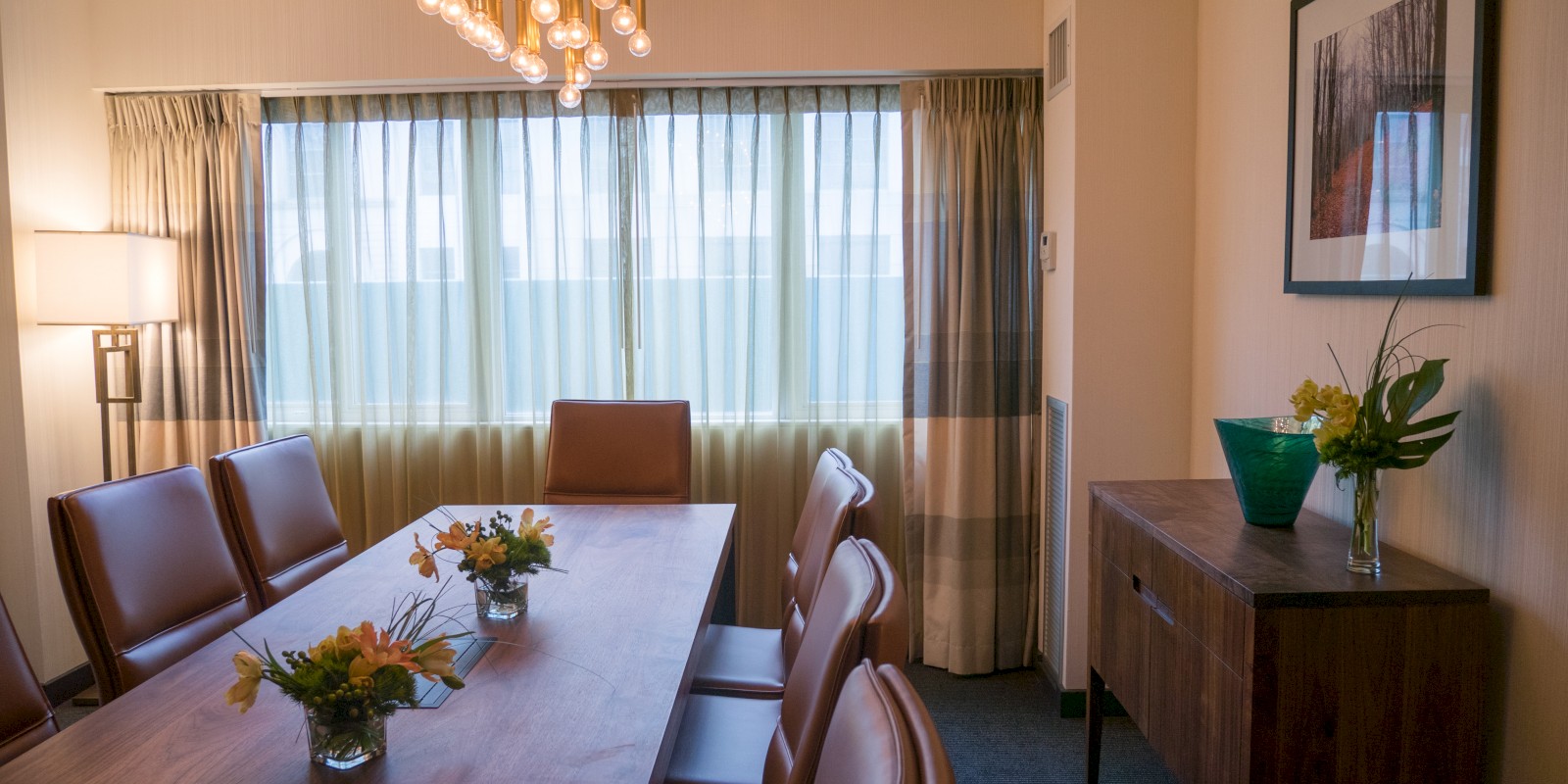 The image shows a conference room with a long table, chairs, a modern light fixture, curtains, and plants on the table and a sideboard.