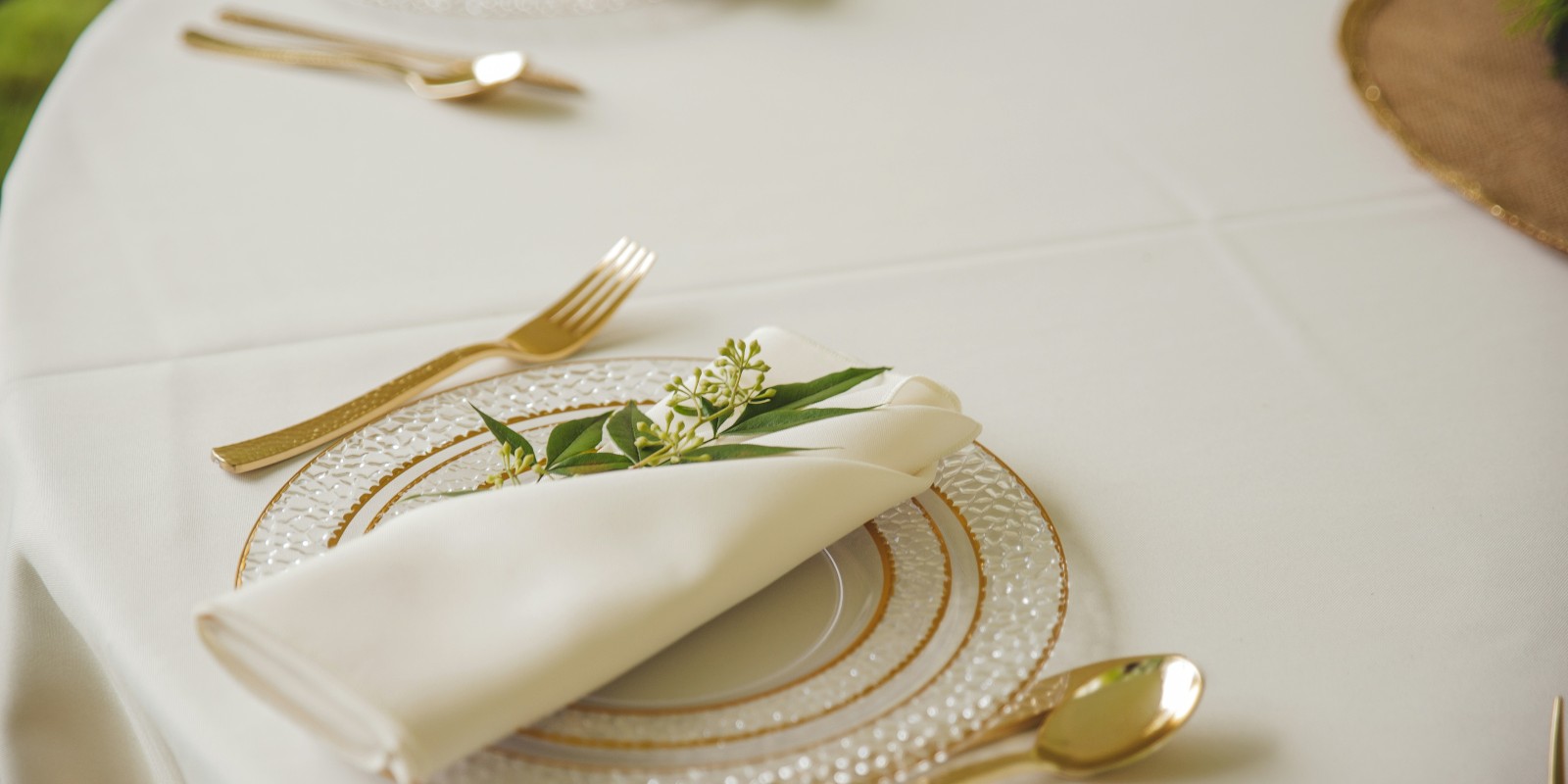 A table setting with gold cutlery, a glass plate, and a white napkin elegantly folded with a small greenery sprig.