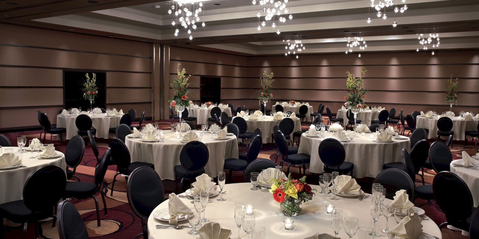 The image shows an elegantly set banquet hall with round tables decorated with flowers, napkins, glassware, and cutlery under modern chandeliers.