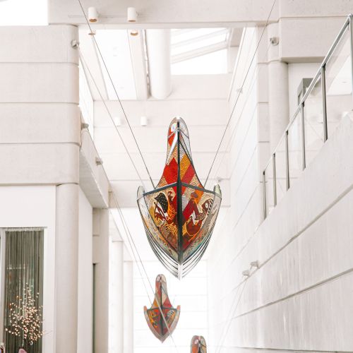 Three colorful boat-like sculptures are suspended from the ceiling in a modern, well-lit atrium with skylights, and white structural elements.