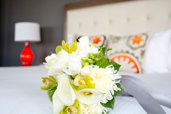 The image shows a bouquet of white and yellow flowers on a bed with decorative pillows and a red lamp in the background.