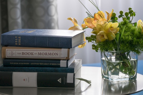 The image shows a stack of religious books including the Bible and the Book of Mormon beside a vase of yellow flowers on a table, ending the sentence.