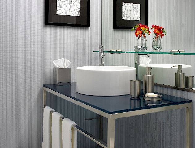 The image shows a modern bathroom sink area with a round basin, mirror, toiletries, towels, and decor including framed art and fresh flowers in a vase.
