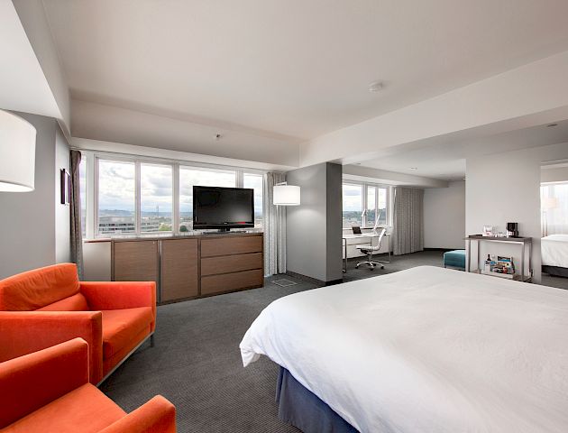 The image shows a modern hotel room with a large bed, an orange chair, a TV on a dresser, a desk with a chair, and large windows.