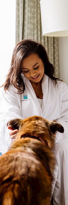 A woman in a white robe is sitting on a blue sofa, smiling and petting a brown dog in a well-lit room.