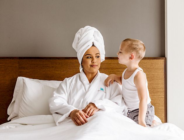 A person in a bathrobe and hair towel sits on a bed with a child in a tank top and shorts standing next to them, both looking content.