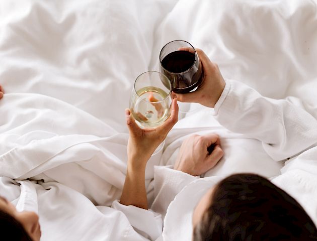 Two people are lying in bed, each holding a glass of wine, one white and one red, appearing to toast each other.
