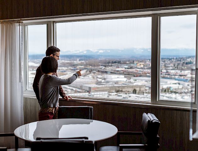 Two people stand by a window, overlooking a cityscape with mountains in the background. One person is pointing outside.