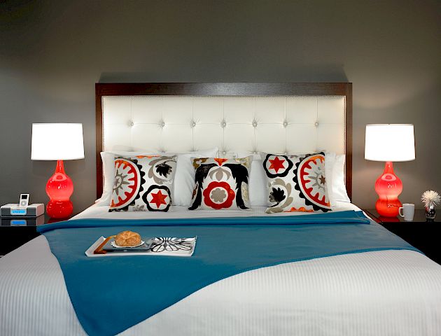 A neatly made bed with vibrant pillows, two red bedside lamps, and a tray with breakfast on a teal blanket at the foot of the bed, ending the sentence.
